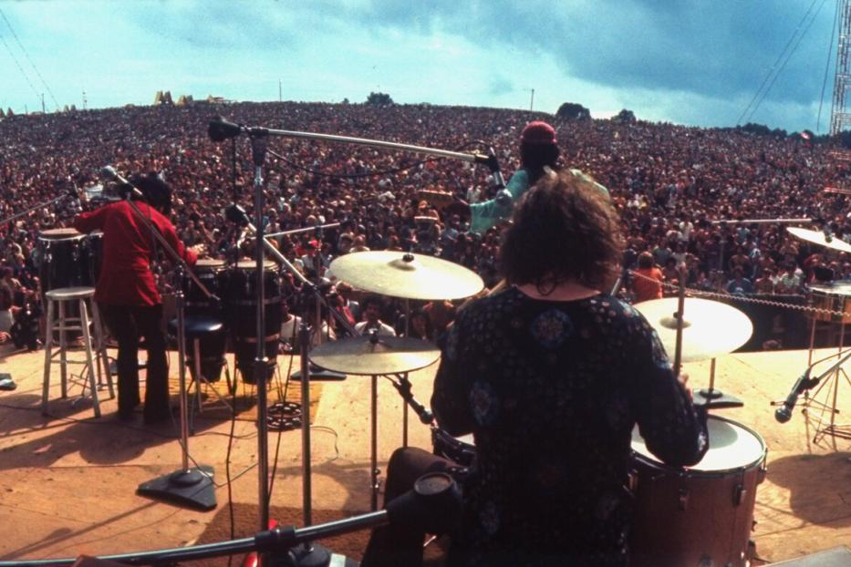 Eine Band auf der Bühne beim Woodstock Festival 1969. Von der Bühne blicken die Musiker inmitten eine Menschenmasse.