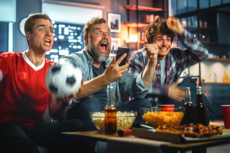 Drei Männer jubeln begeistert beim gemeinsamen Fußballschauen auf dem Sofa mit Snacks und Getränken.
