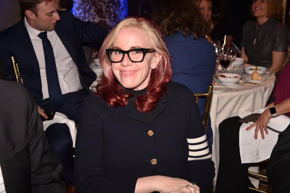 Eine Frau mit blond-roten Haaren und Brille sitzt an einem Tisch.