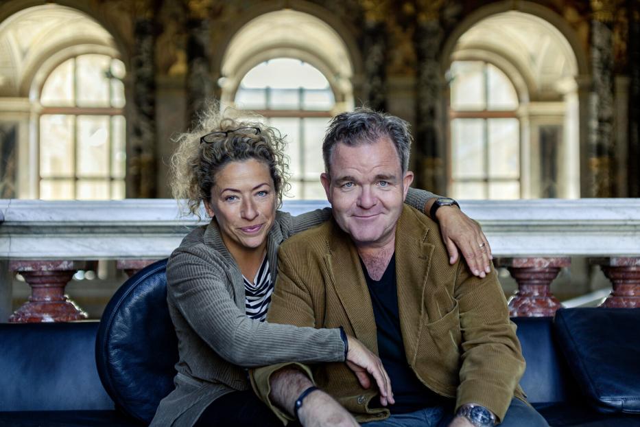 Carolin Pienkos und Cornelius Obonya sitzen auf einer Bank. Sie hat den Arm um ihn geschlungen, beide schauen sehr freundlich.