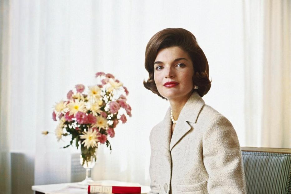 Ein Porträt von Jackie Kennedy in einem Tweed-Kostüm neben einem Blumenarrangement.