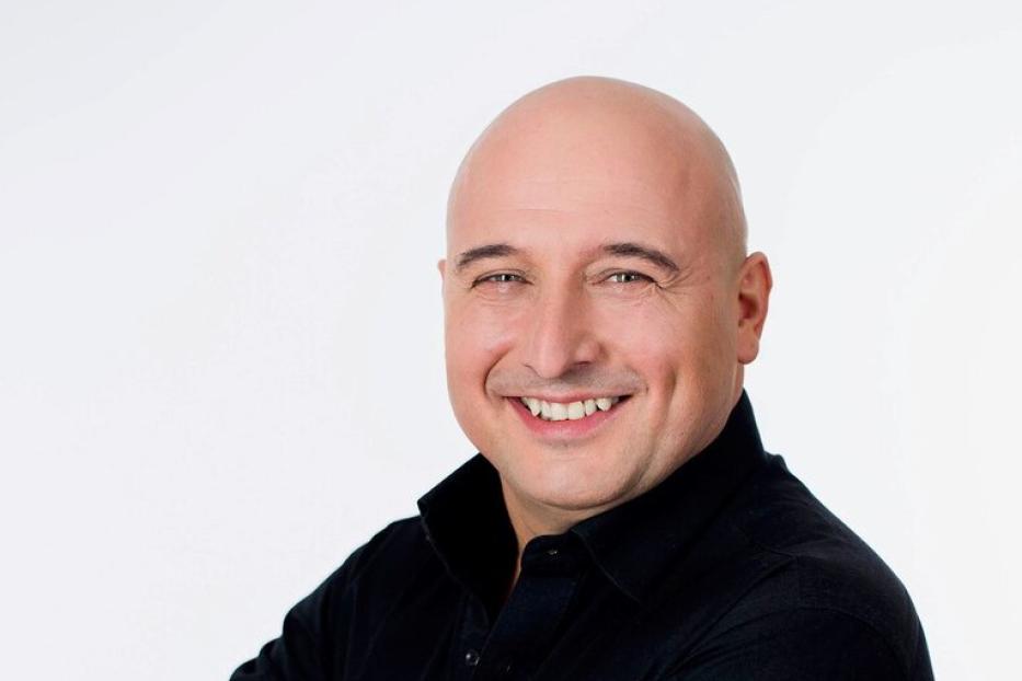 Ein lächelnder, glatzköpfiger Mann mit verschränkten Armen vor einem weißen Hintergrund.
