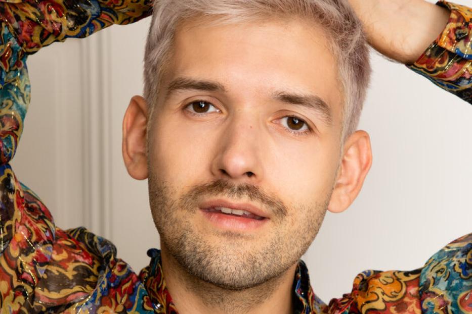 Ein Mann mit blond gefärbten Haaren und einem bunten Hemd posiert vor einer weißen Wand.