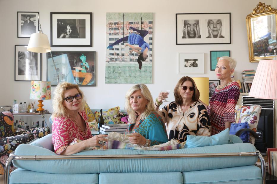 Vier Frauen sitzen auf einem Sofa und halten Gläser in einem Wohnzimmer mit Kunst an den Wänden.