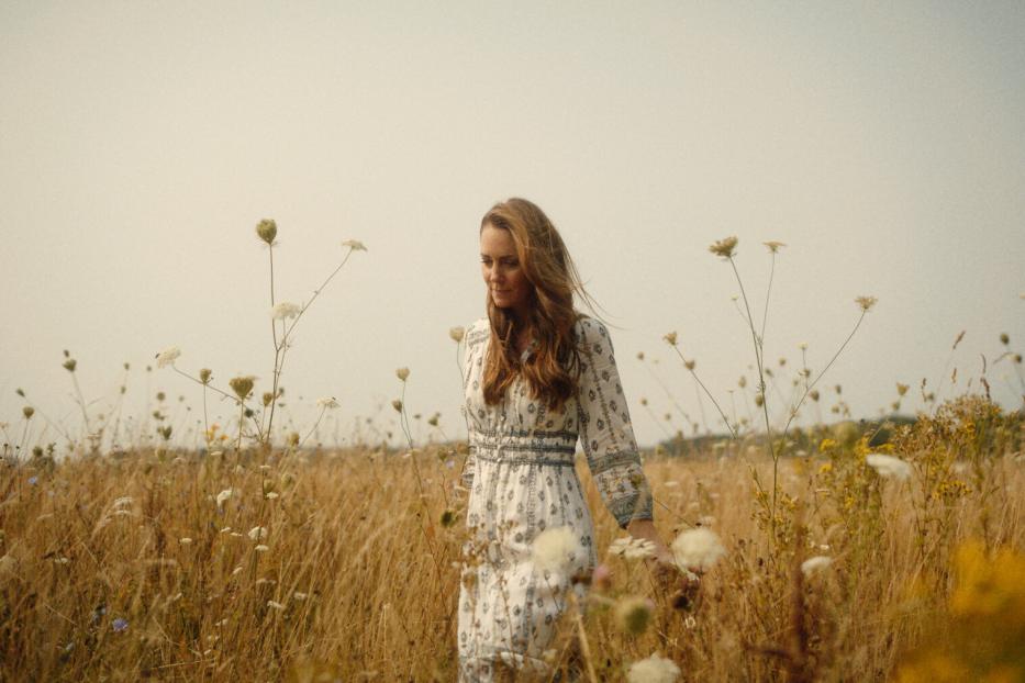 Eine Frau mit langem Haar geht durch ein Feld mit hohem Gras und Wildblumen.