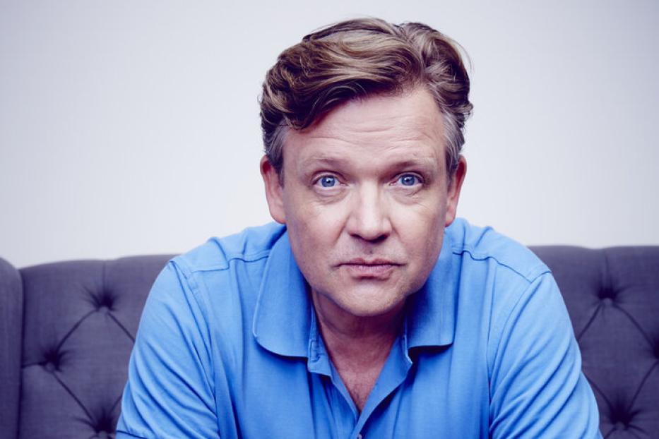 Ein Mann mit blauem Poloshirt sitzt auf einem grauen Sofa.