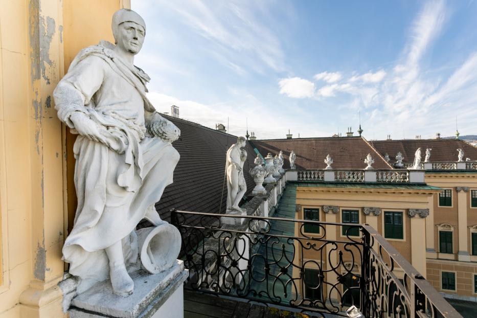 Steinfiguren schauen beim Balkon unterhalb der großen Uhr von oben auf den Ehrenhof des Schlosses Schönbrunn herab. Vor dem Balkon befindet sich ein Schmiedeisengitter, vor dem Dach eine steinerne Brüstung.