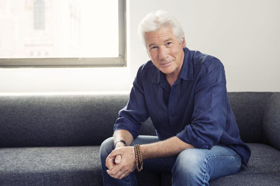 Richard Gere sitzt auf einer grauen Couch vor einem Fenster.
