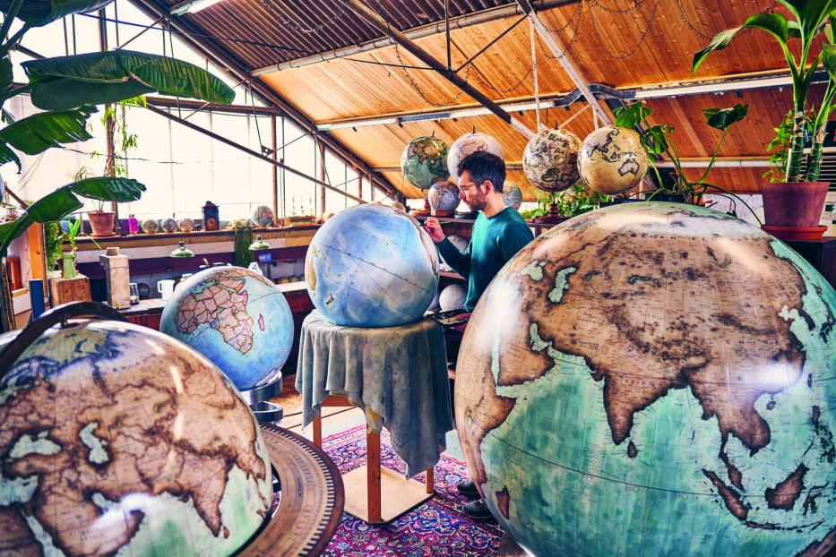 Ein Mann bemalt in einem Atelier einen Globus, umgeben von weiteren Globen unterschiedlicher Größe.