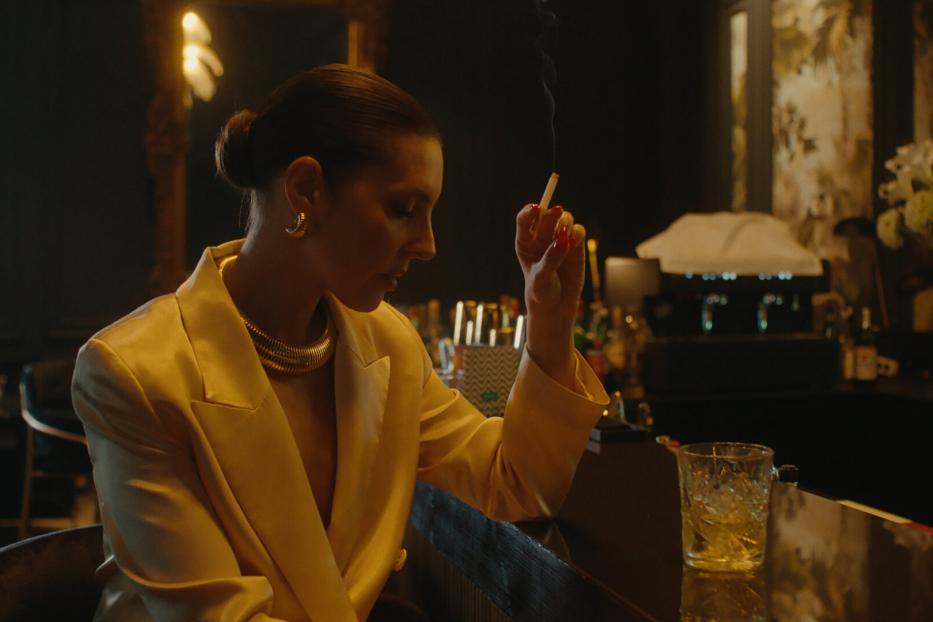 Eine Frau im gelben Blazer raucht an einer Bar.