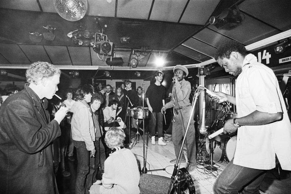Auf einer kleinen Bühne in einer niedrigen Disco spielt eine Band, da Publikum raucht, sitzt und steht herum.