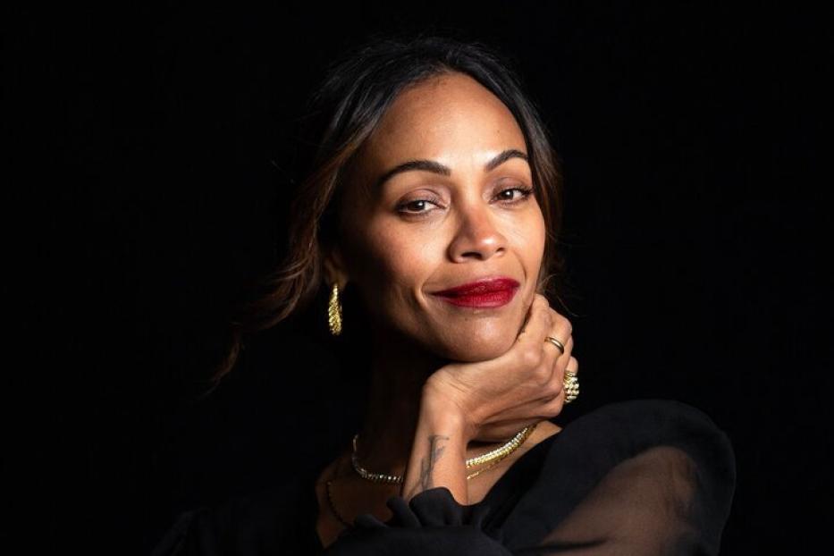 Zoe Saldana posiert vor einem dunklen Hintergrund in einem schwarzen, transparenten Oberteil.