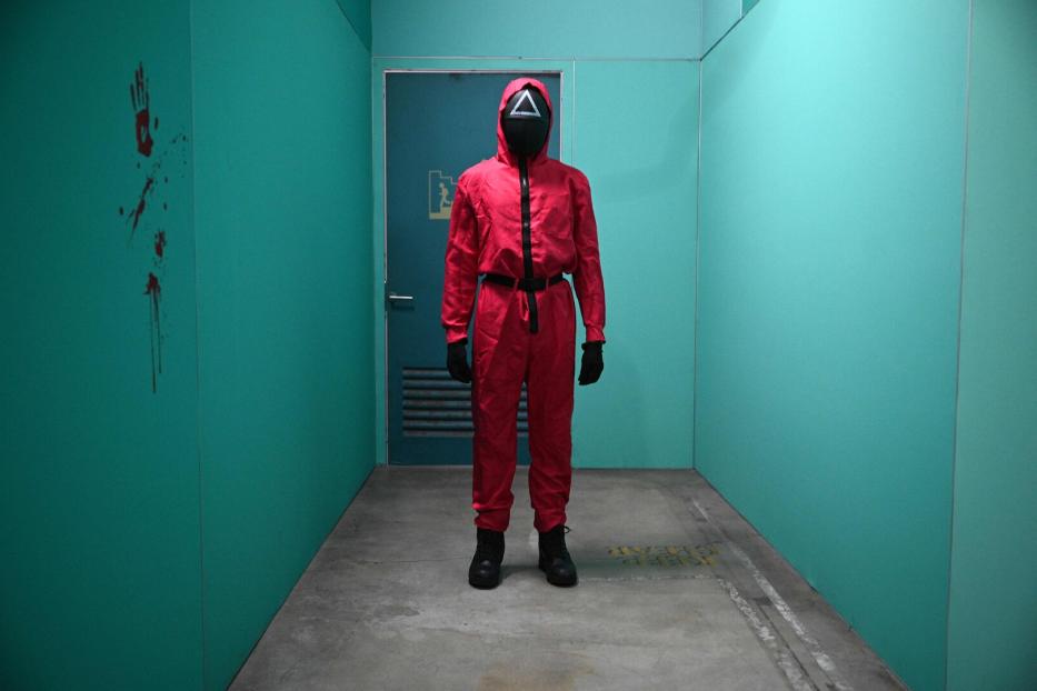 Eine Person in rotem Overall und Maske mit Dreieck steht in einem grünen Gang.