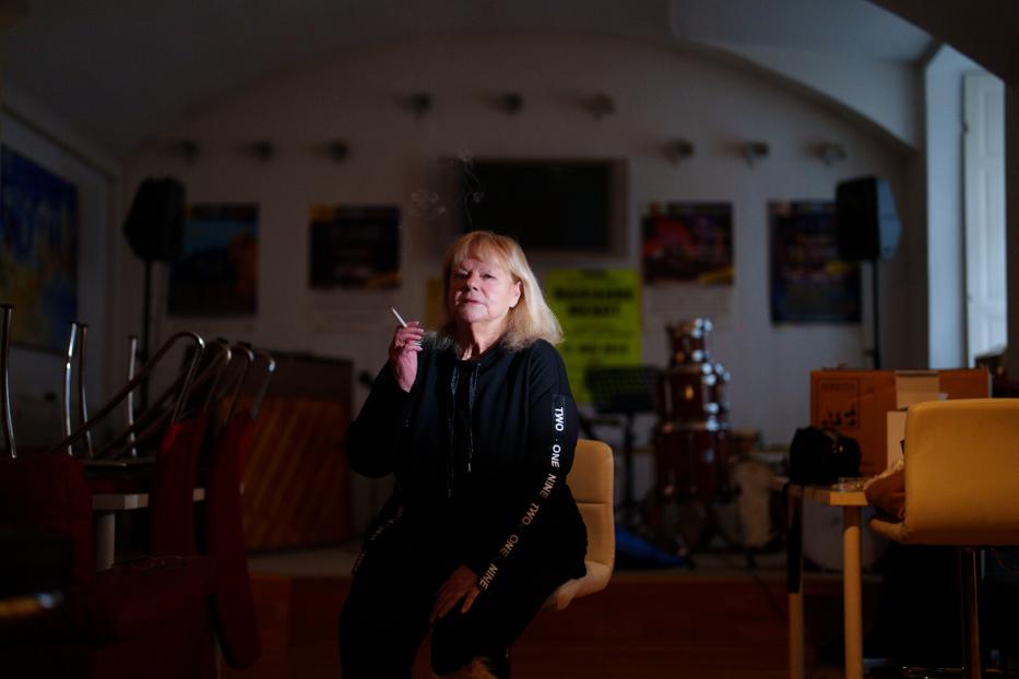 Eine Frau mit blonder Bob-Frisur raucht eine Zigarette in einem Raum mit Musikinstrumenten.