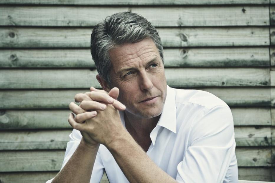 Hugh Grant sitzt an einem Tisch und blickt nachdenklich zur Seite.