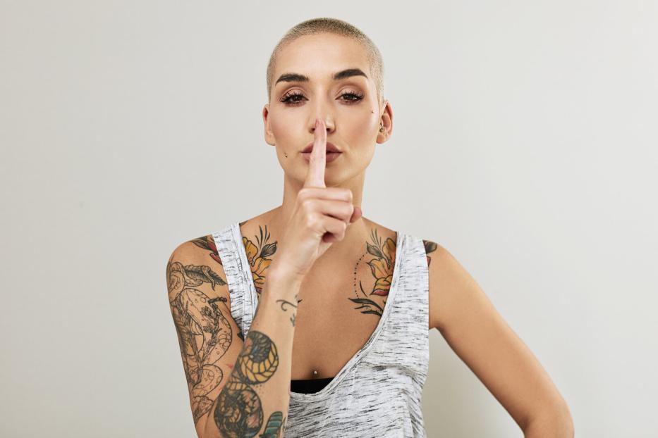 Eine junge Frau mit Tattoos hält den Finger vor den Mund.