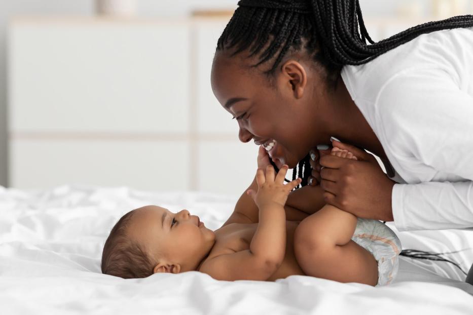 Eine lächelnde Mutter spielt mit ihrem Baby auf einem weißen Bett.