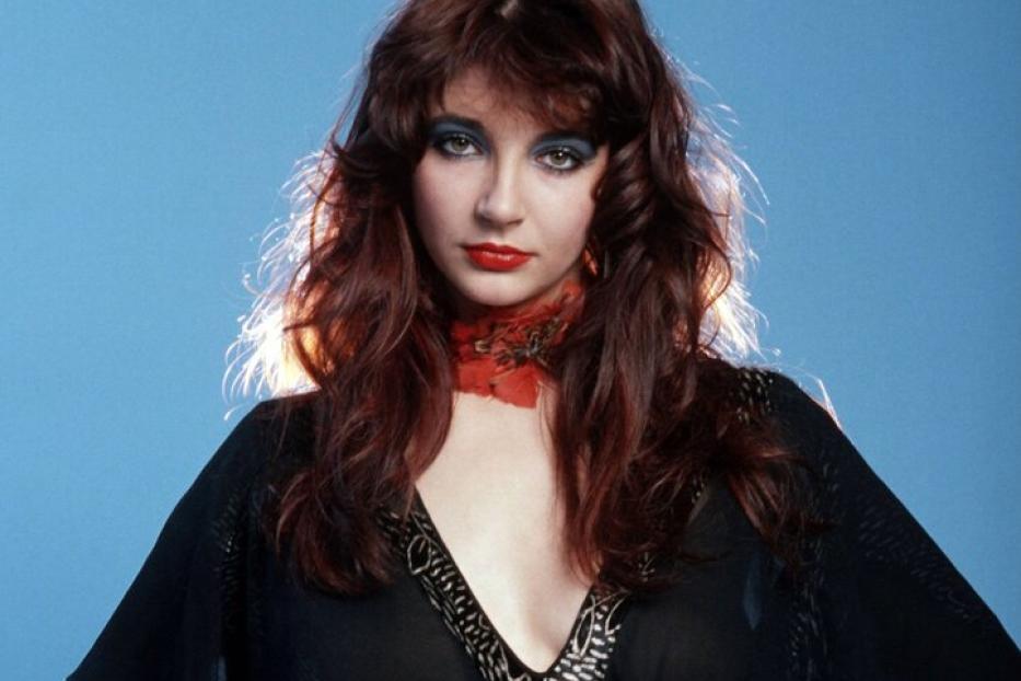 Kate Bush posiert vor einem blauen Hintergrund mit den Händen auf den Hüften.