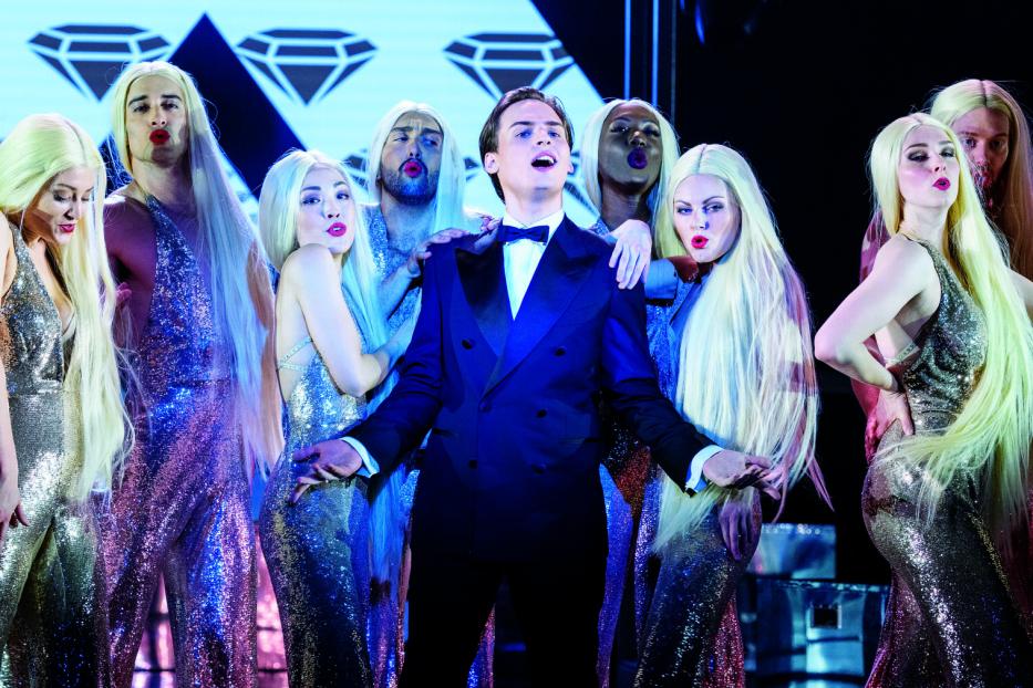 Ein Mann im Anzug singt, umgeben von Personen in glitzernden Kleidern und blonden Perücken.