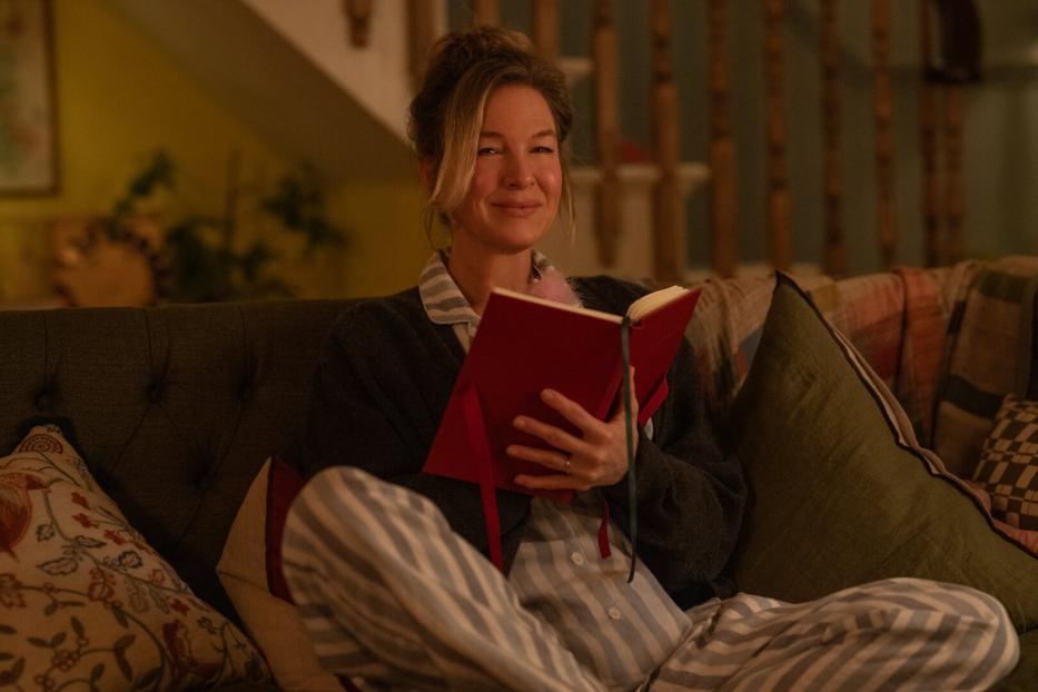 Renée Zellweger sitzt im Schlafanzug auf einem Sofa und liest in einem roten Buch.