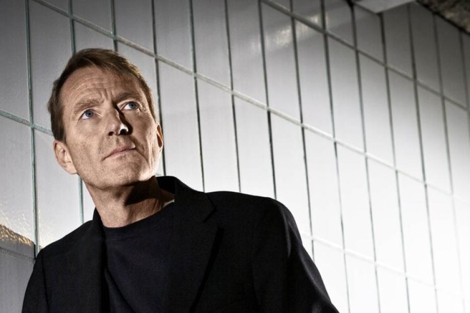 Ein Mann in einem schwarzen Anzug lehnt an einer gefliesten Wand und schaut nach oben.