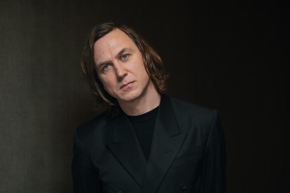Porträt von Jarvis Cocker in einem schwarzen Anzug vor einem dunklen Hintergrund.
