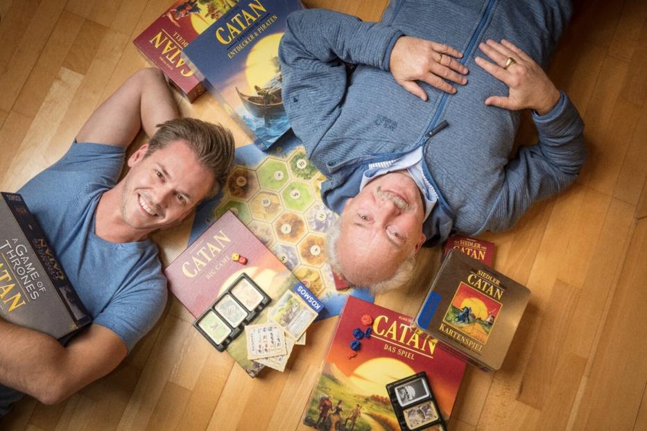 Zwei Männer liegen auf dem Boden, umgeben von verschiedenen „Catan“-Spielen.