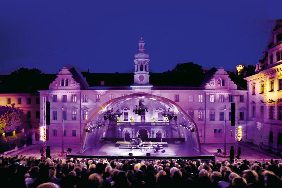 Eine Open-Air-Veranstaltung mit einer Bühne vor einem historischen Gebäude zieht viele Zuschauer an.