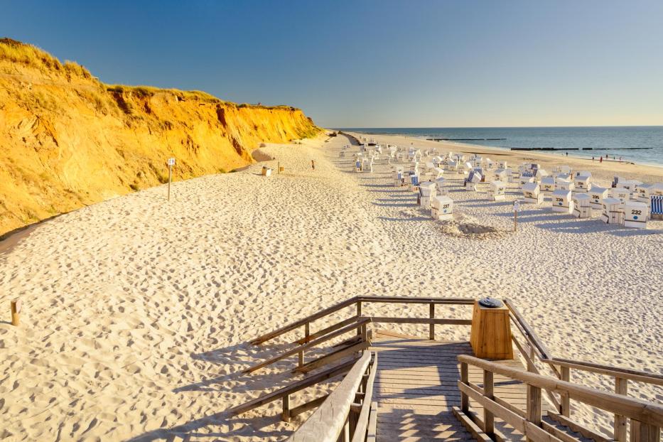 Ein weißer Strand mit Strandkörben und einer Klippe hintendran.