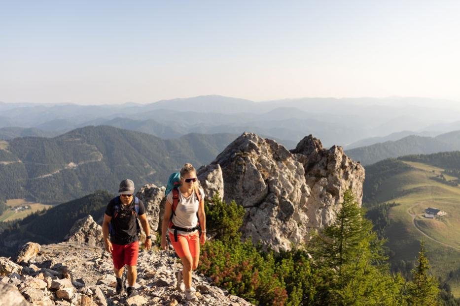 Zwei Wanderer steigen auf einem felsigen Pfad mit Blick auf eine Berglandschaft auf.