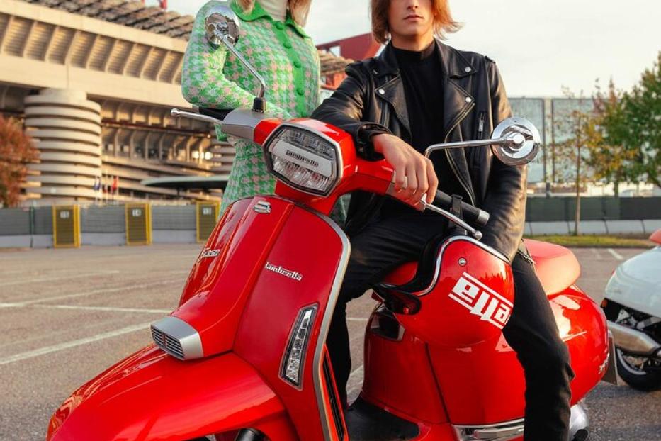 Ein Mann und eine Frau posieren auf einem roten Lambretta-Roller vor einem Stadion.