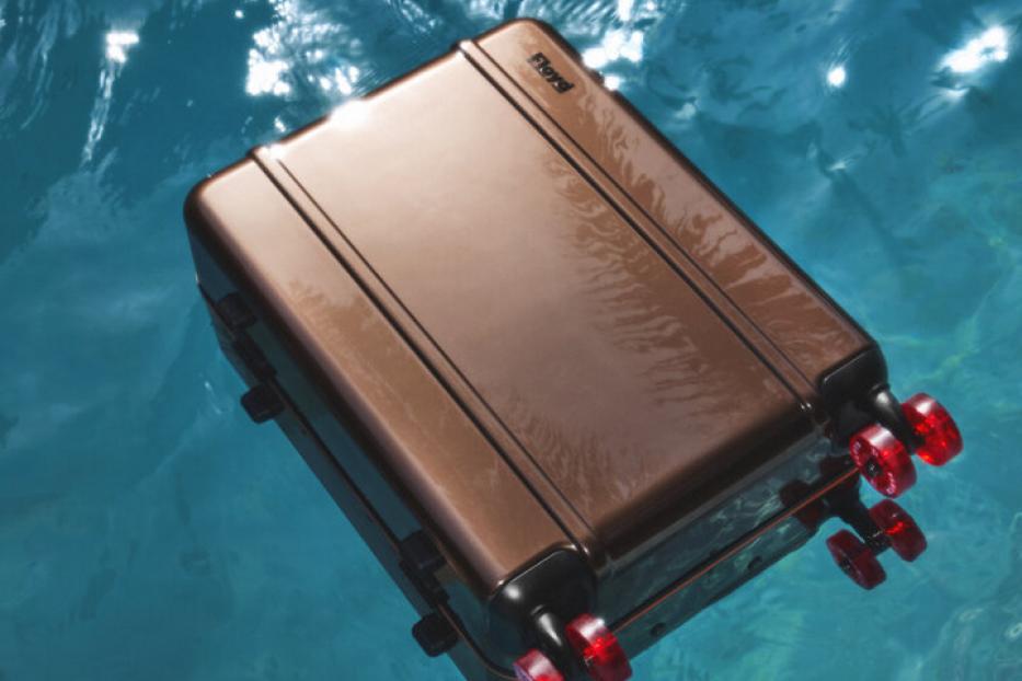 Ein bronzefarbener Rollkoffer treibt auf dem türkisfarbenen Wasser.
