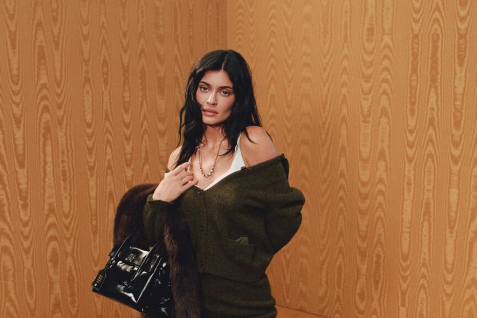 Kylie Jenner posiert für Miu Miu in einem grünen Off-Shoulder-Cardigan und einem passenden Rock dazu. Außerdem trägt sie Kniestrümpfe und schwarze Schuhe zum Schnürren. Ihre Accessoires sind eine goldene Kette, eine kleine Ledertasche und ein brauner Pelzmantel.