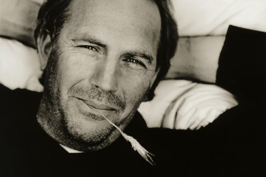 Ein Schwarzweißporträt von Kevin Costner mit einem Grashalm im Mund.