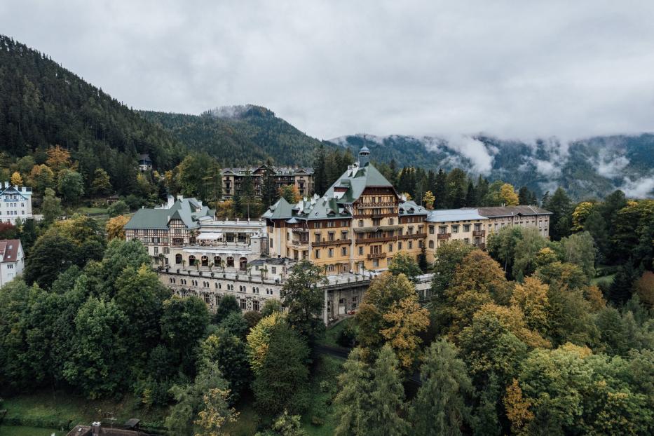 Das große Südbahnhotel mit seinen verschiedenen Trakten und großem Balkon. Es thront auf einem bewaldeten Hang. Im Hintergrund erheben sich Berge, Nebel lichtet sich.