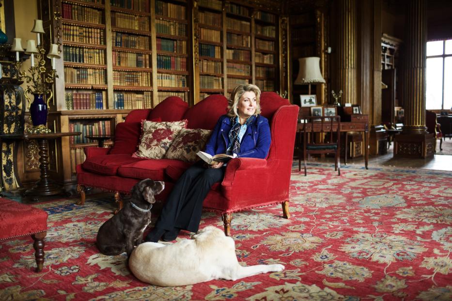 Eine Frau liest auf einem roten Sofa in Highclere Castle, während zwei Hunde auf dem Teppich liegen.