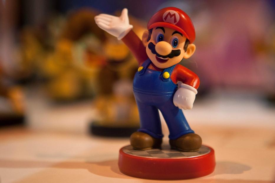 Eine Mario-Figur steht auf einem Schachbrettmuster-Sockel und winkt.