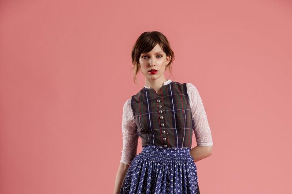 Eine Frau in einem Dirndl mit blauem Rock und karierter Weste vor einem rosa Hintergrund.