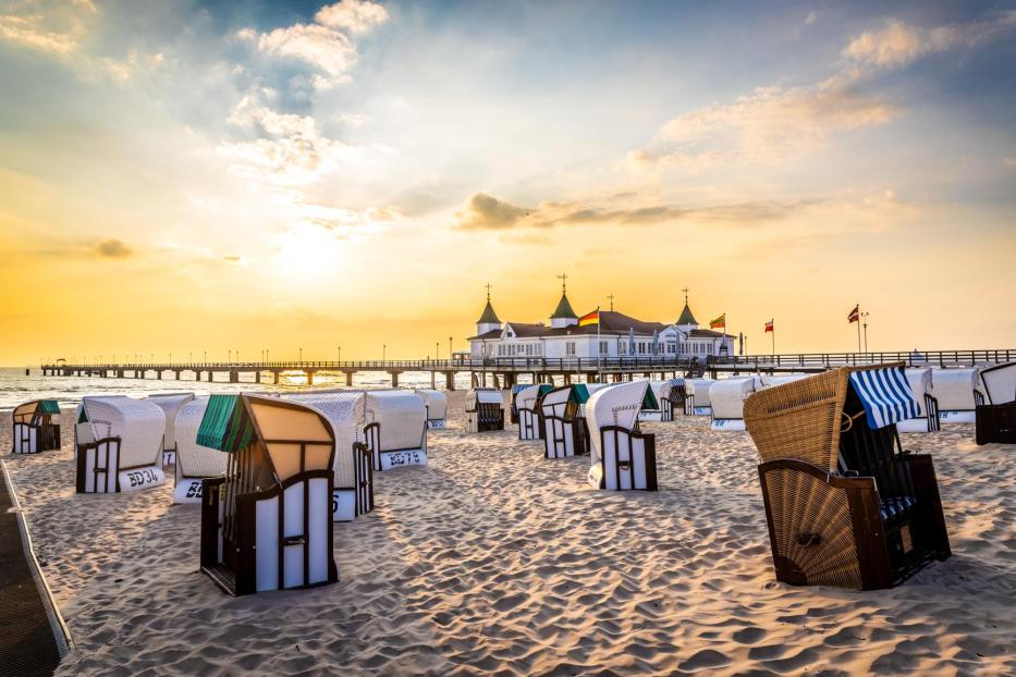 Strand mit mehreren Strandkörben in verschiedenen Farben und einer Seebrücke mit Gebäude im Hintergrund bei Sonnenuntergang.