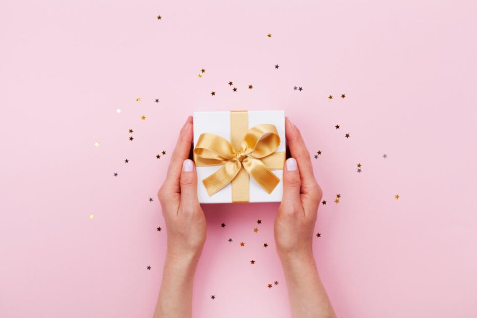 Zwei Hände halten eine weiße Geschenkbox mit goldener Schleife auf rosa Hintergrund mit kleinen Sternkonfetti.