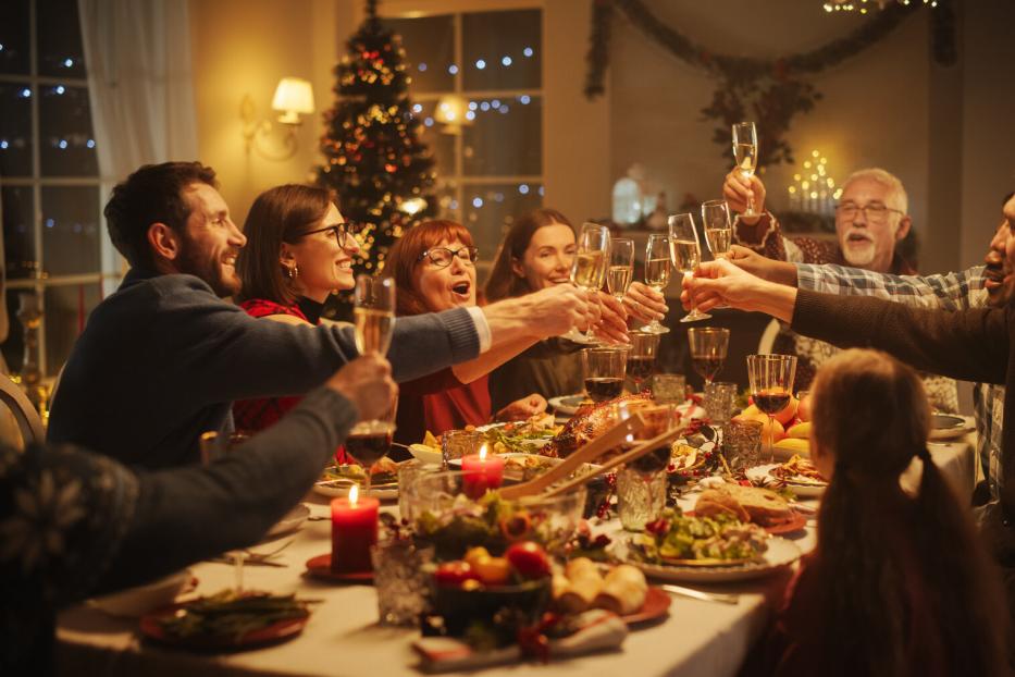 Mehrere Menschen stoßen an einem festlich gedeckten Tisch mit Sektgläsern an, im Hintergrund steht ein geschmückter Weihnachtsbaum.