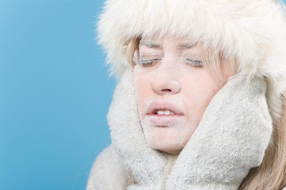 Eine Frau mit frostbedecktem Gesicht trägt eine Pelzmütze und Handschuhe vor blauem Hintergrund.