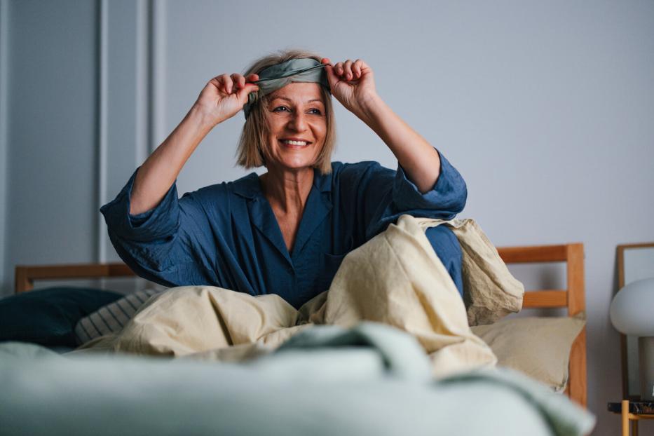 Eine lächelnde Frau im blauen Pyjama sitzt im Bett und zieht sich eine Schlafmaske vom Kopf.