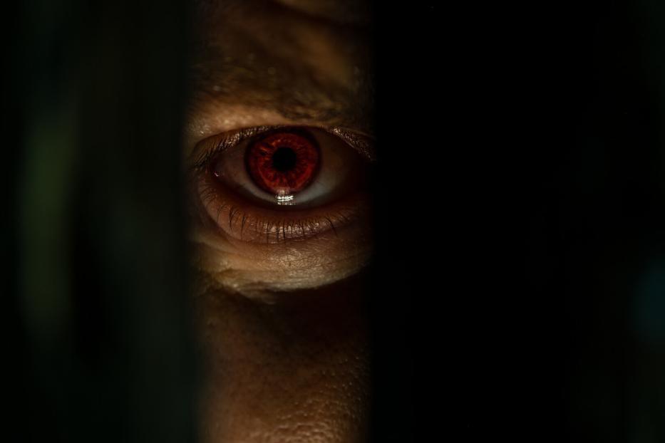 Ein rotes menschliches Auge blickt durch einen schmalen dunklen Spalt.