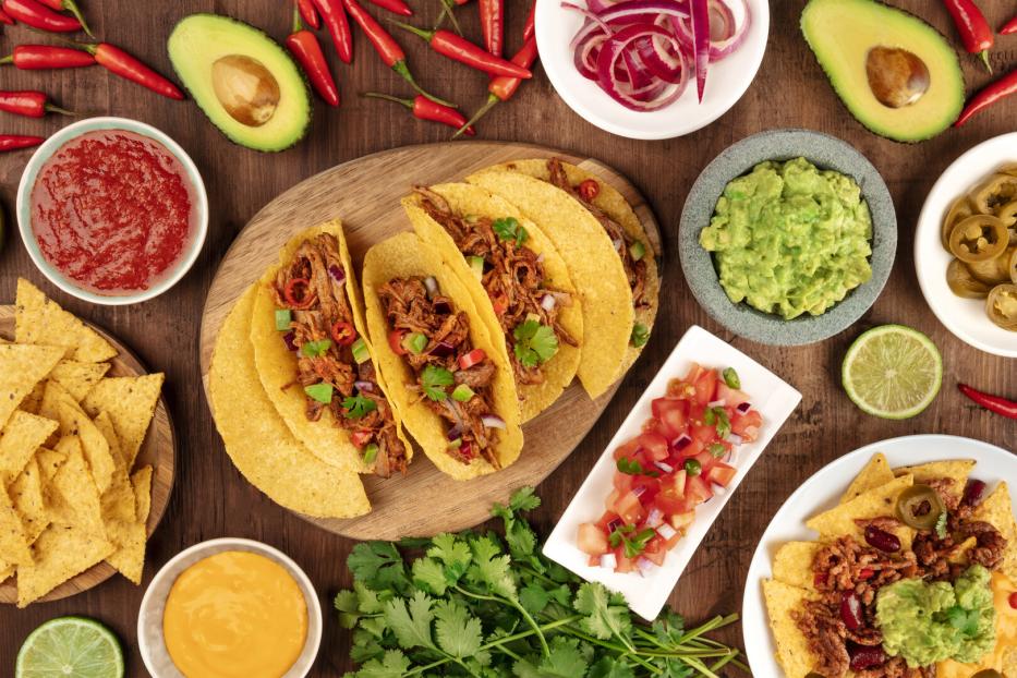 Mexikanisches Essen mit Tacos, Tortilla-Chips, Guacamole, Salsa, Käse, Limetten, Chili und Jalapeños auf Holztisch.