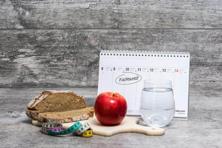 Ein Apfel, ein Glas Wasser, Brot, ein Maßband und ein Kalender mit dem Wort „Fastenzeit“ auf einem Holztisch.
