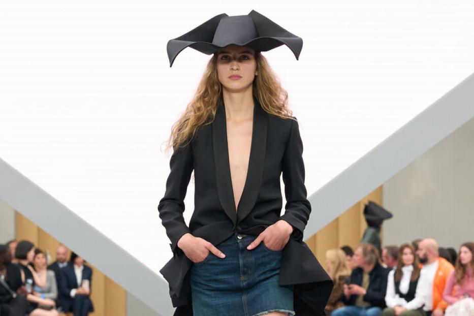 Eine Frau trägt einen tief ausgeschnittenen schwarzen Blazer, einen Jeansrock und einen auffälligen schwarzen Hut auf einem Laufsteg.
