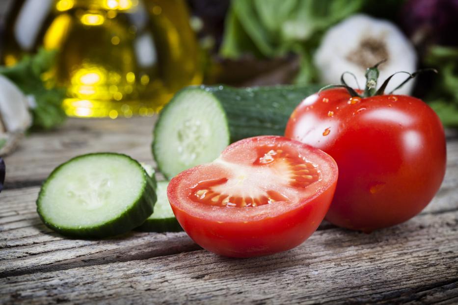 Eine ganze Tomate, eine halbierte Tomate und mehrere Gurkenscheiben liegen auf einem Holztisch.