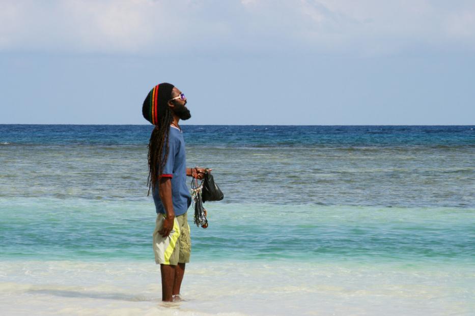 Ein Mann mit Dreadlocks und Rasta-Mütze steht im flachen türkisfarbenen Meer und hält Schmuckstücke in der Hand.