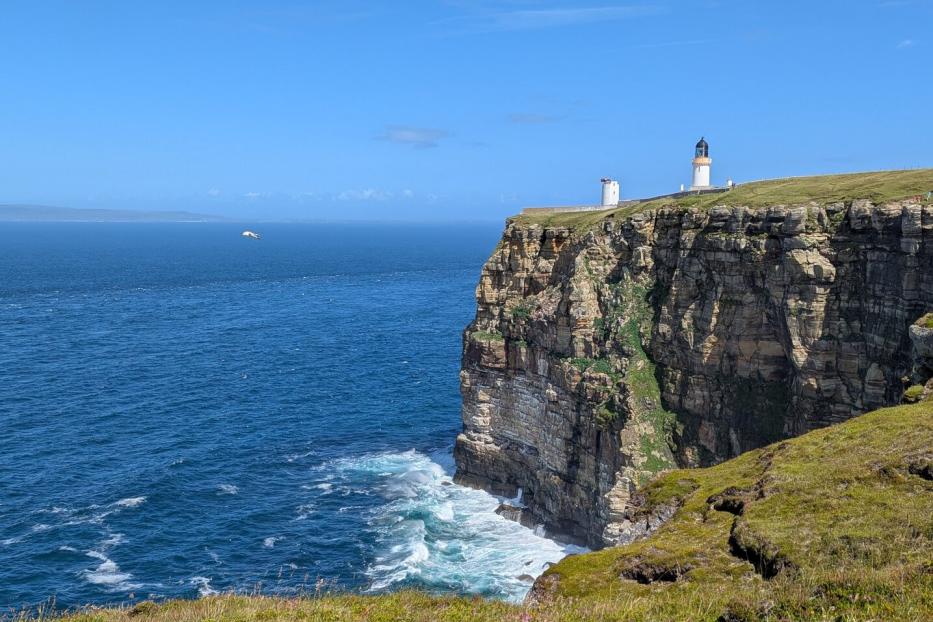Steilküste am Meer mit Leuchtturm auf der Klippe und Wellenbrandung darunter.
